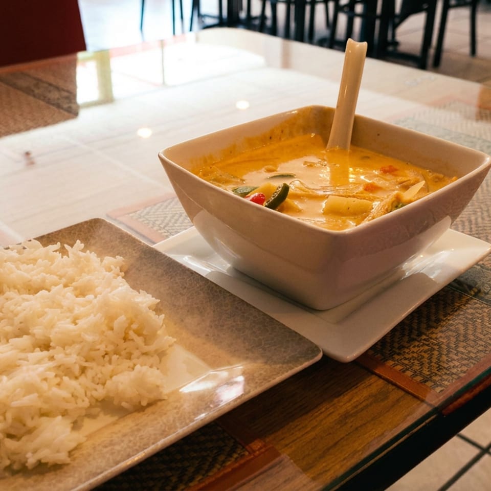 Square bowl of creamy orange curry with vegetables and spoon, served alongside white rice on a restaurant table.