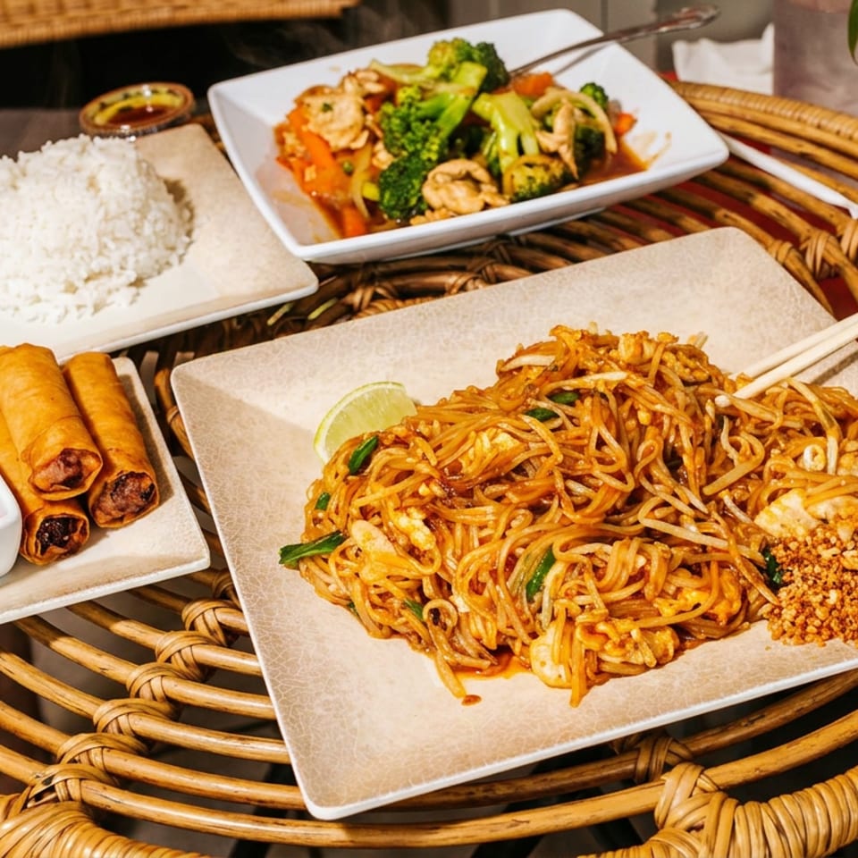 Asian meal featuring stir-fried noodles, vegetable chicken stir-fry, steamed rice, spring rolls, dipping sauce, and chopsticks on a wicker table.