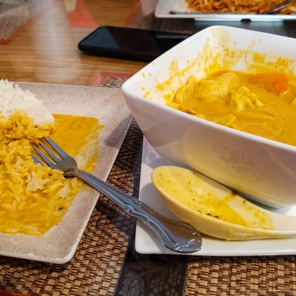 Square bowl of yellow curry with vegetables beside rice, a spoon, fork, and partially visible restaurant dishes on a wooden table.